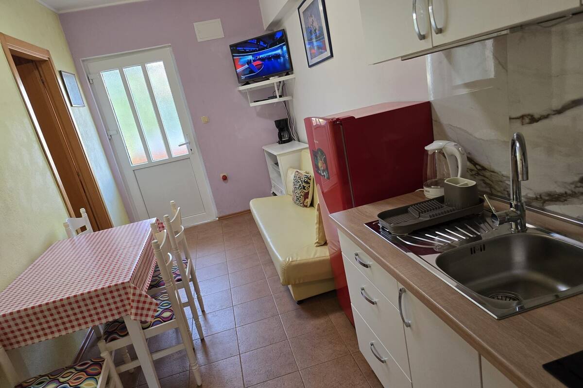 A compact kitchen with a sink, a small refrigerator, a television mounted above the counter, and a dining area with a table and chairs.