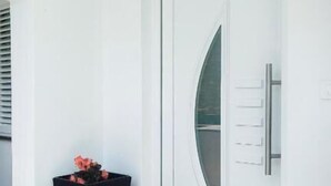 A white door with a glass panel and a handle, a black planter with flowers, and a wall-mounted light fixture. - House Solis (Rab)