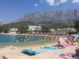 A beach with clear blue water, people swimming and sunbathing, and mountains in the background.