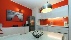 A modern kitchen with white cabinets, a red accent wall, a stainless steel refrigerator, and a dining table with a unique black centerpiece. - Apartment Mandria (Opatija)