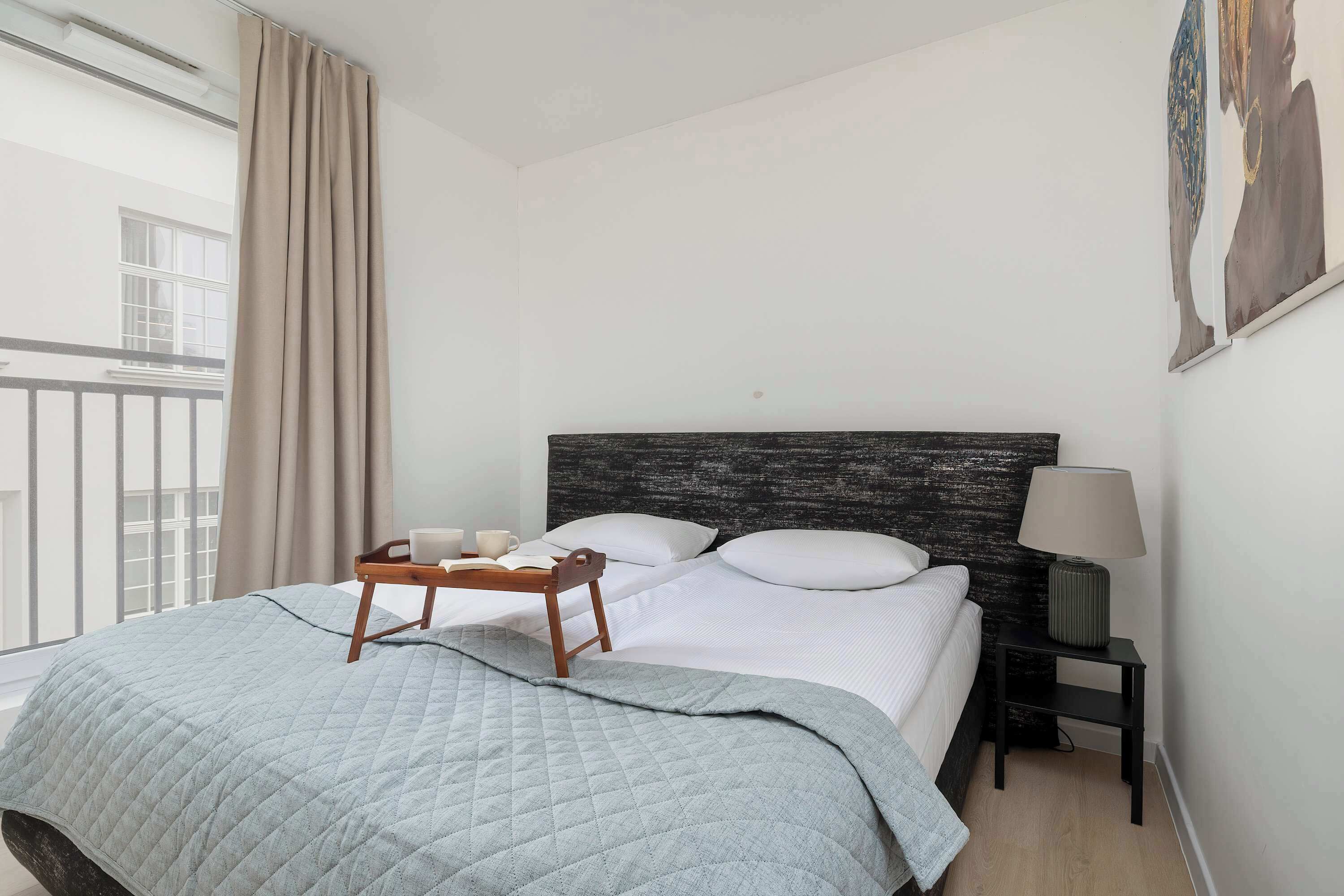 Un lit bien fait, recouvert d’une couverture grise, une table de chevet avec une lampe et une fenêtre avec des rideaux.