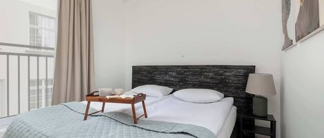 Un lit bien fait, recouvert d’une couverture grise, une table de chevet avec une lampe et une fenêtre avec des rideaux.