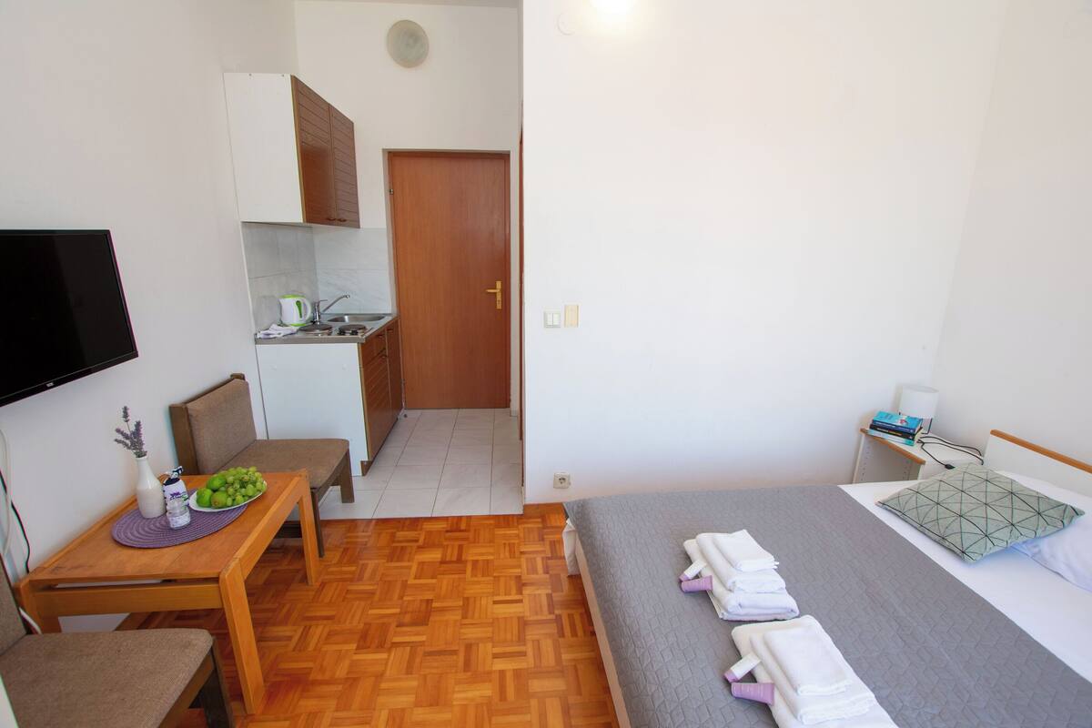 Une chambre d’hôtel compacte comprenant un lit, une petite table avec un vase et un panier de fruits, une chaise, un coin cuisine et un téléviseur fixé au mur.
