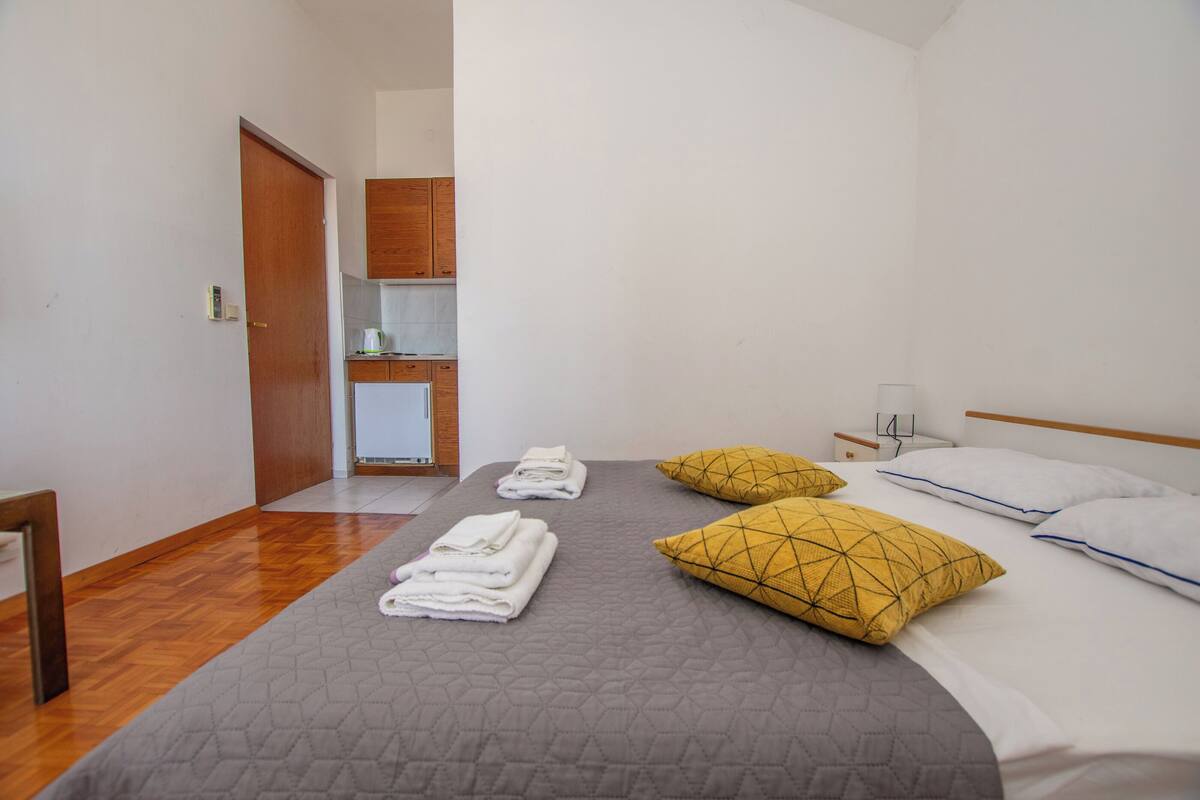 Une chambre d’hôtel avec un lit, une table de chevet, une lampe, une porte en bois et, au fond, une petite cuisine.