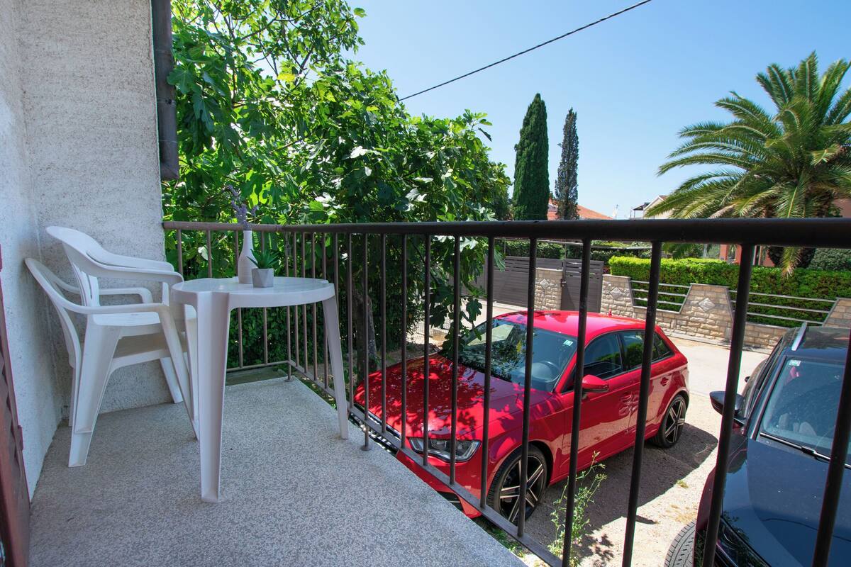 Un balcon avec une table et des chaises, une voiture rouge garée devant, et une vue sur la verdure et un bâtiment en arrière-plan.