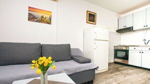 Una sala de estar con un sofá gris, una mesita con un jarrón de flores amarillas, un refrigerador blanco y una zona de cocina con gabinetes blancos y una estufa.