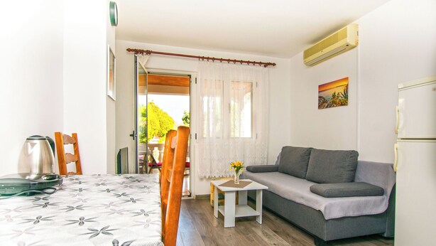 Una habitación con una mesa de comedor, un sofá, una mesita con un florero, un refrigerador y una ventana con cortinas.