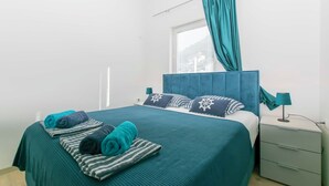 A neatly made bed with teal bedding, striped pillows, and rolled towels. There is a window with blinds and a teal curtain. A bedside table with a lamp and a drawer is visible. - Apartments Beach House Blace (Slivno)