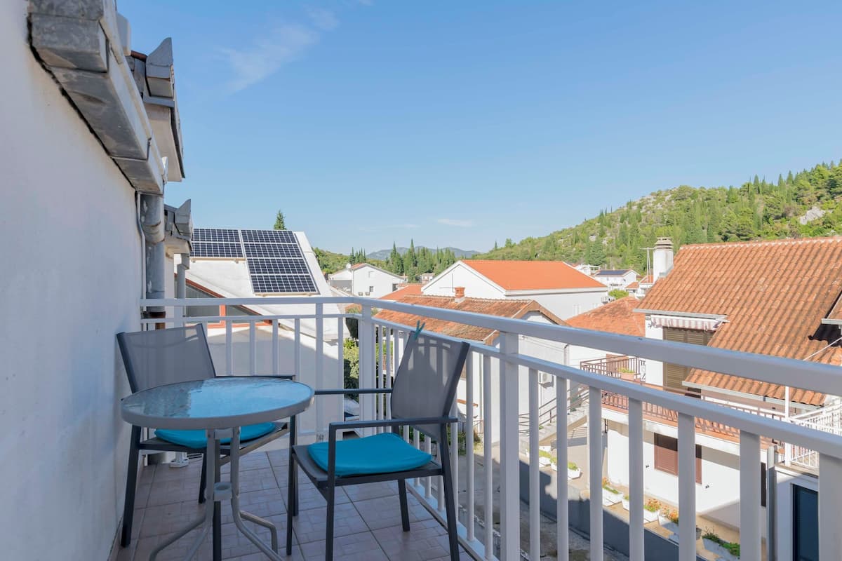 A balcony with a table and chairs, solar panels, and a view of neighboring buildings and hills.