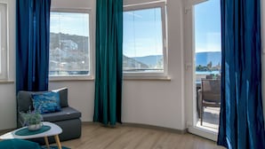 A modern room with a grey sofa, a small round table, and a view of the city through large windows with blue curtains. - Apartments Beach House Blace (Slivno)