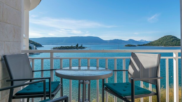 A balcony with two chairs and a small table, overlooking a body of water with boats and distant islands. - Apartments Beach House Blace (Slivno)