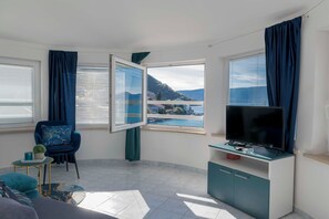 Uma sala de estar moderna com televisão, uma poltrona azul, uma mesinha com uma planta e vista para um corpo de água e montanhas através de grandes janelas.