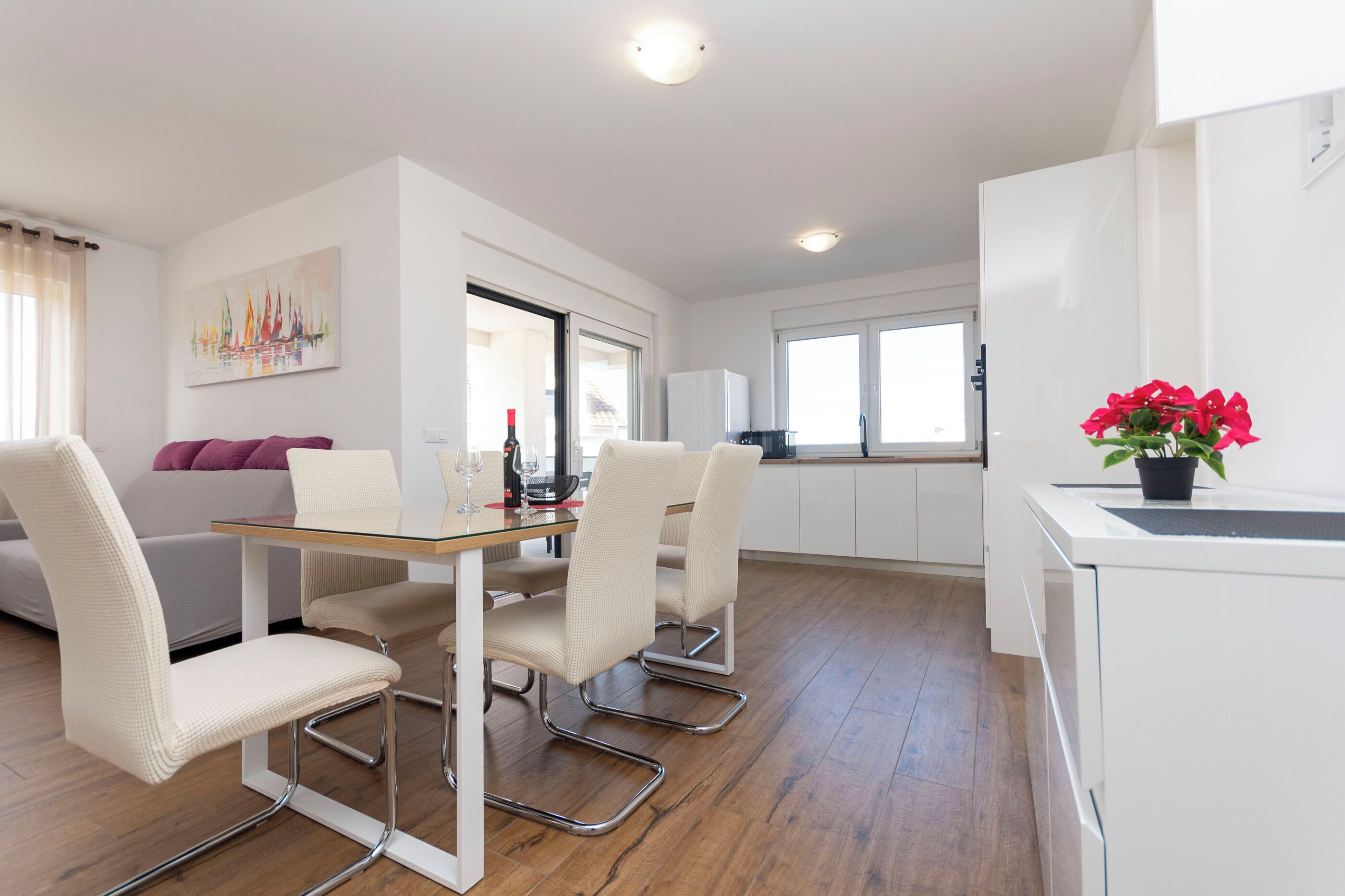 Una cocina moderna con armarios blancos, un comedor con mesa de vidrio y una sala de estar con sofá y un cuadro.