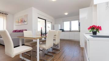 Una cocina moderna con armarios blancos, un comedor con mesa de vidrio y una sala de estar con sofá y un cuadro.