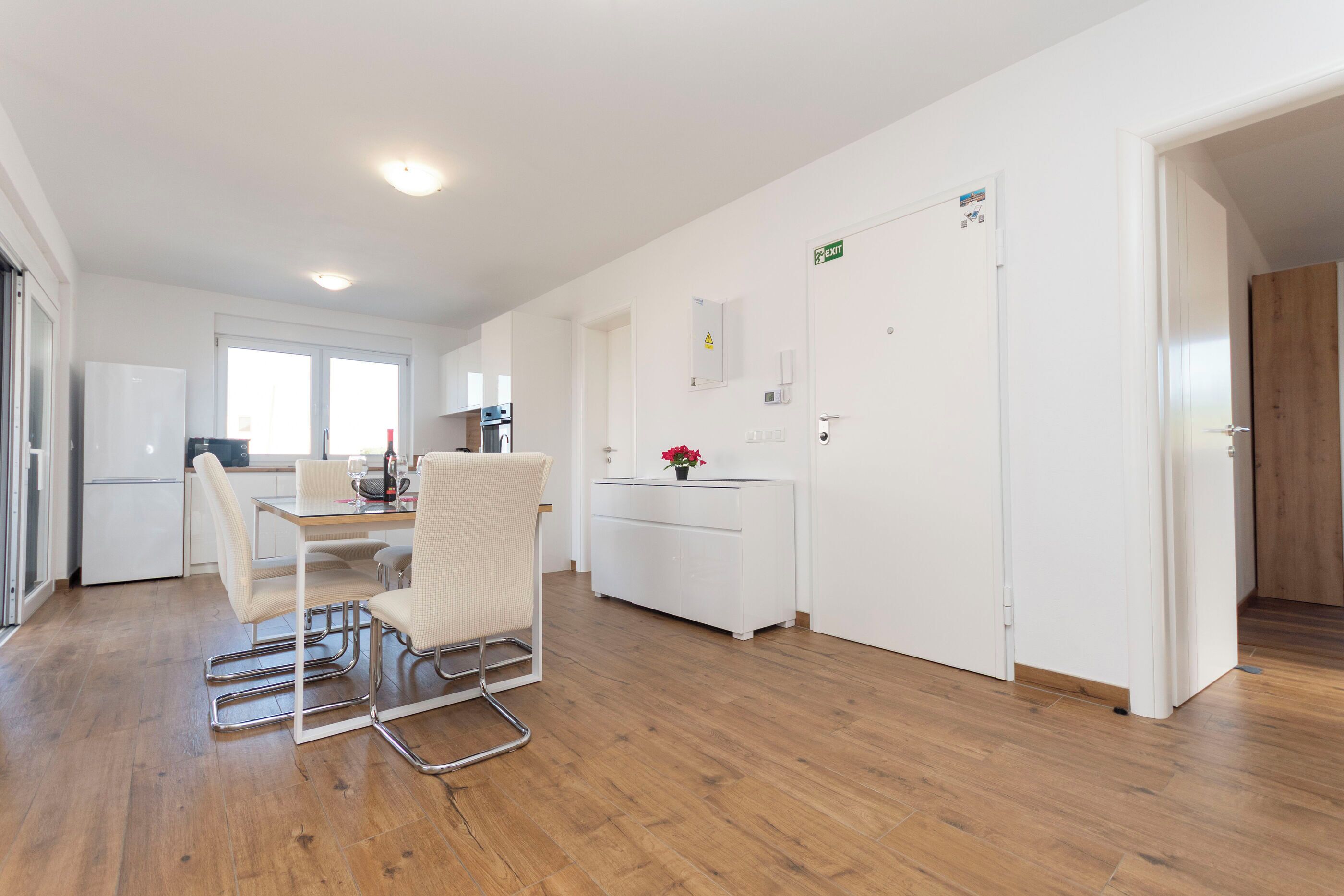 A modern kitchen with white appliances, a dining area with a table and chairs, and a hallway with an exit sign.