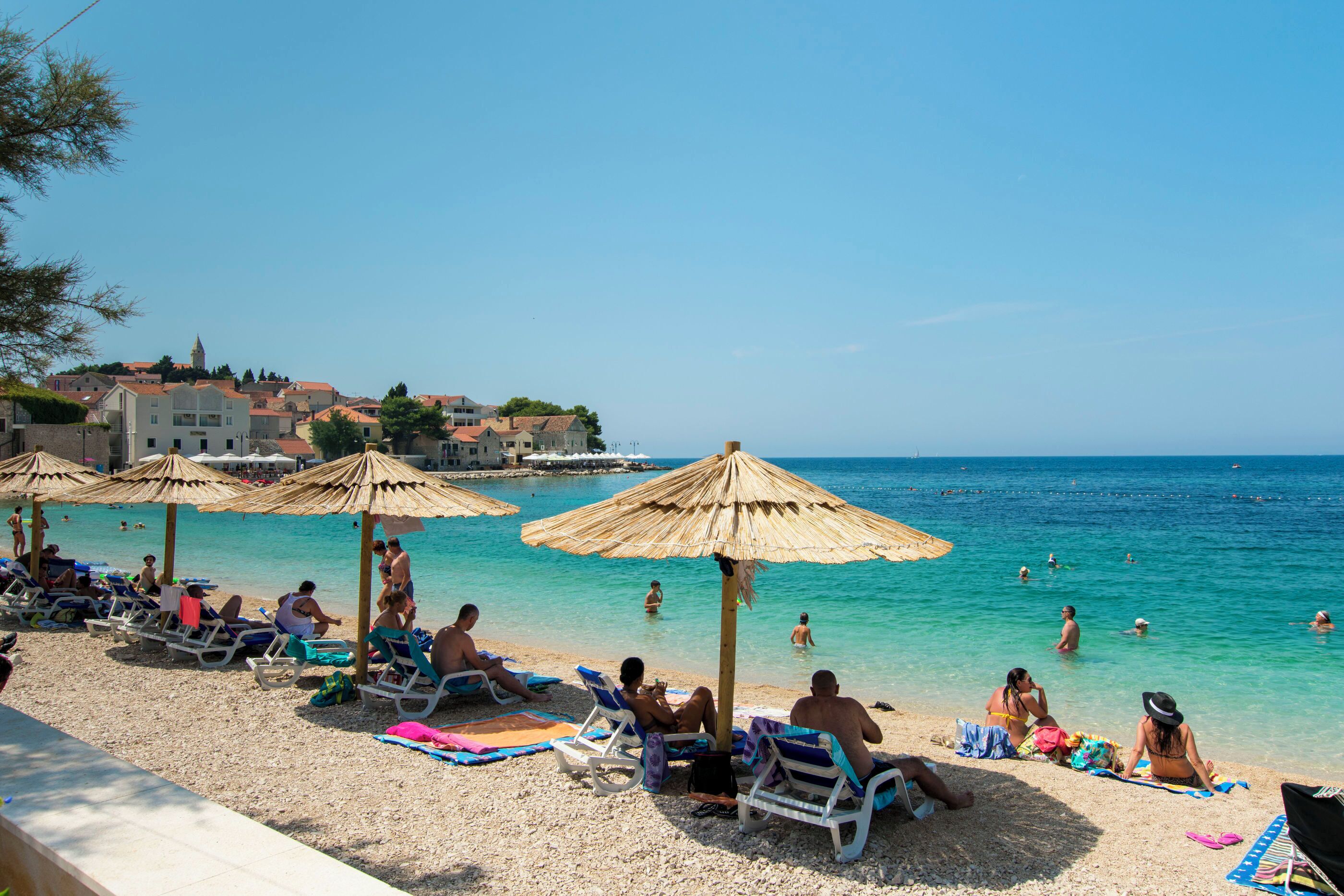 Ein Strand mit Liegestühlen und Sonnenschirmen, klares blaues Wasser und Gebäude im Hintergrund.