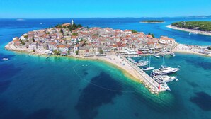 An aerial view of a coastal town with a dense cluster of buildings, a marina with boats, and clear blue waters. - Apartments Pajo Plus (Primosten)