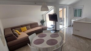 A living room with a brown sofa, a round table with placemats, a TV, and a sliding glass door leading to a balcony. - Apartments Pajo Plus (Primosten)