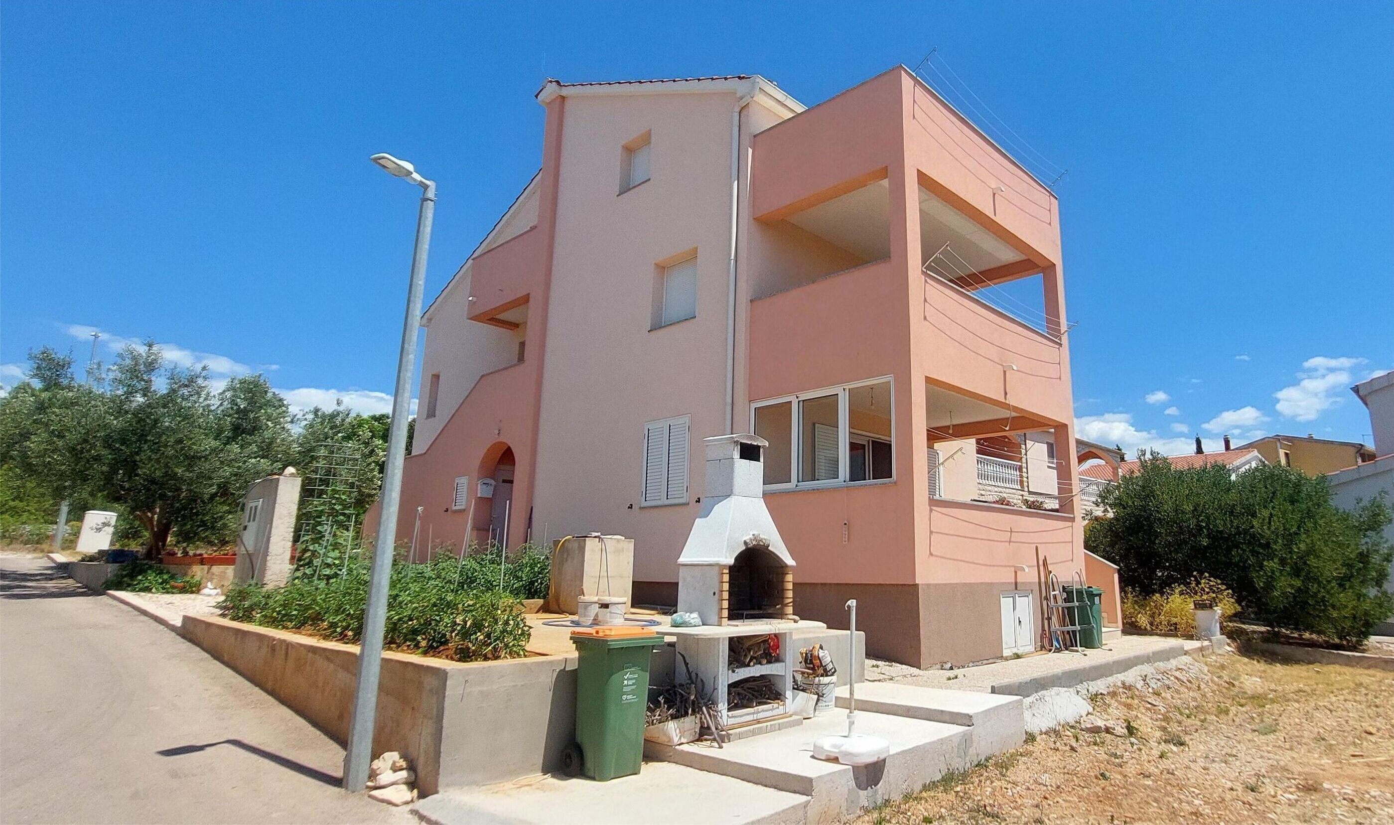 A two-story residential building with a central chimney, a balcony, and a small outdoor area with a trash bin.