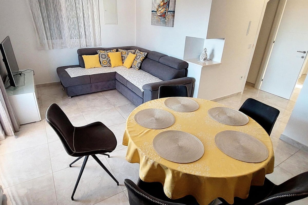 Una sala de estar moderna con sofá, una mesa de comedor con individuales y un televisor sobre un mueble.