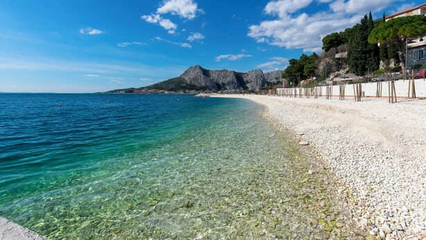 A pebble beach with clear water, a mountain in the background, and buildings along the shore. - Apartment Protrka (Omis)