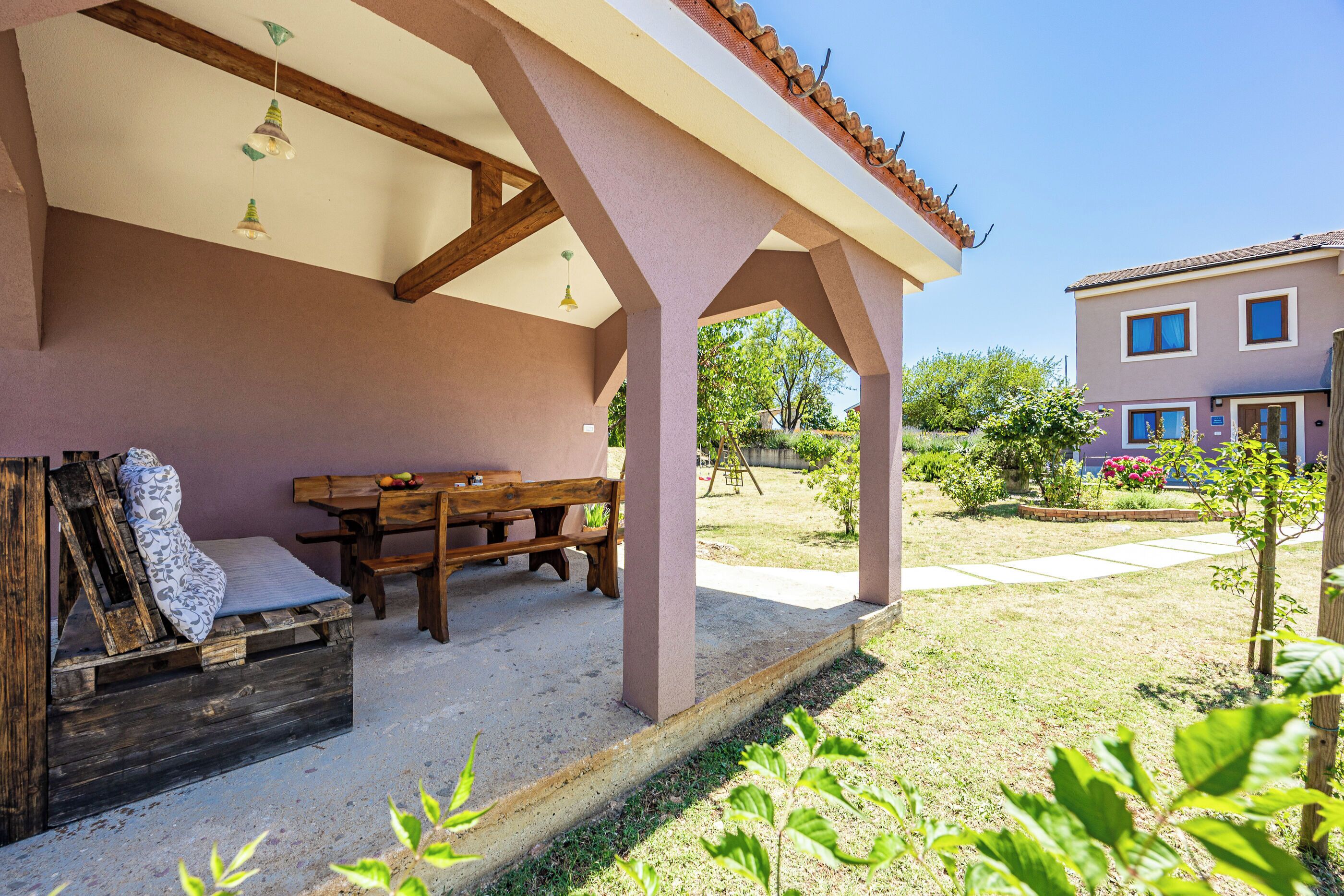 A covered outdoor seating area with a wooden table and benches, a wooden crate with a cushion, and a garden view.
