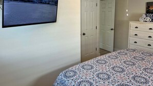 Un dormitorio con cama, cómoda, televisor montado en la pared y una puerta que da a otra habitación.