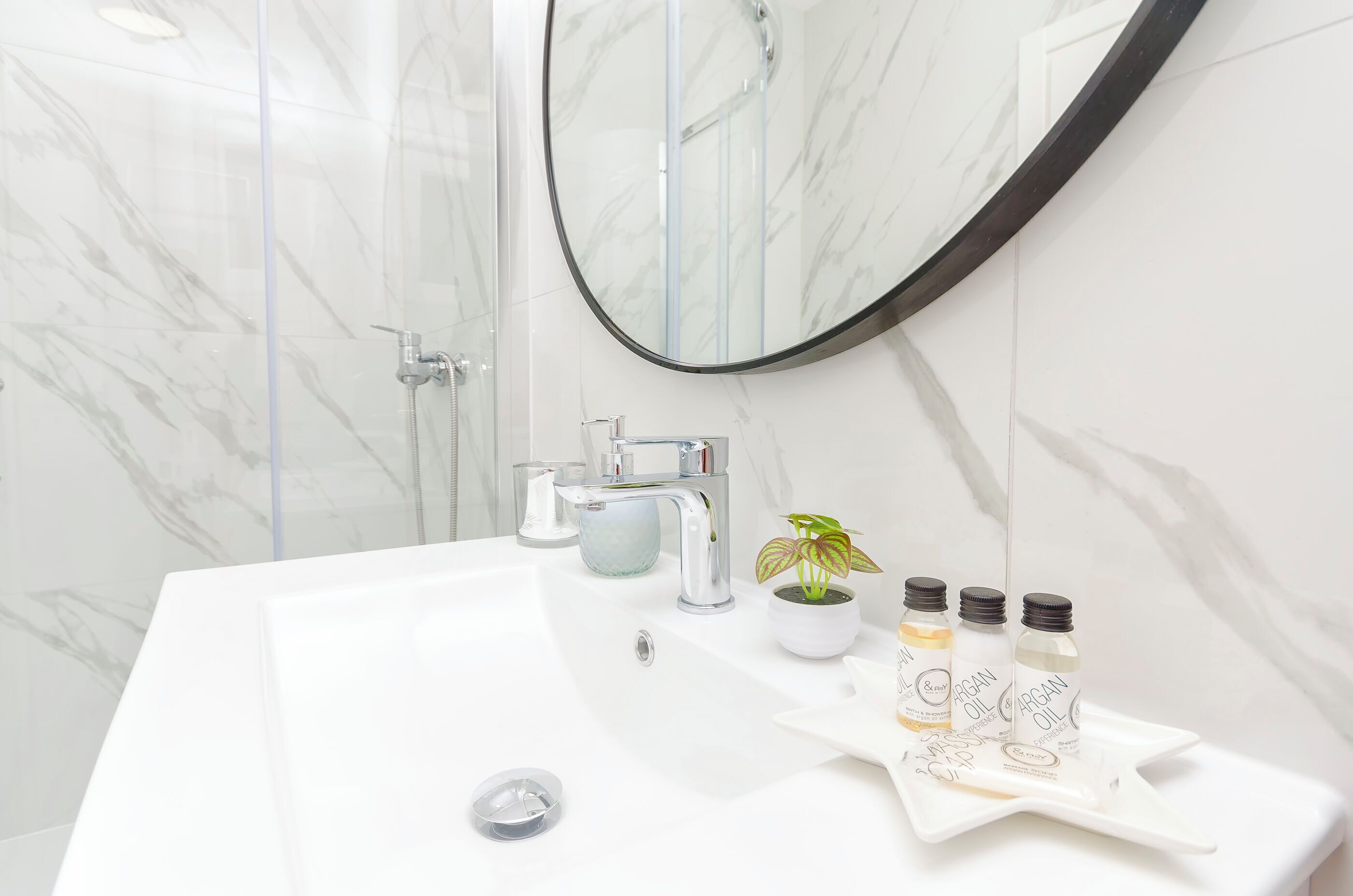 A bathroom with a white sink, a mirror, and a shower. There are three bottles of argan oil on the sink.