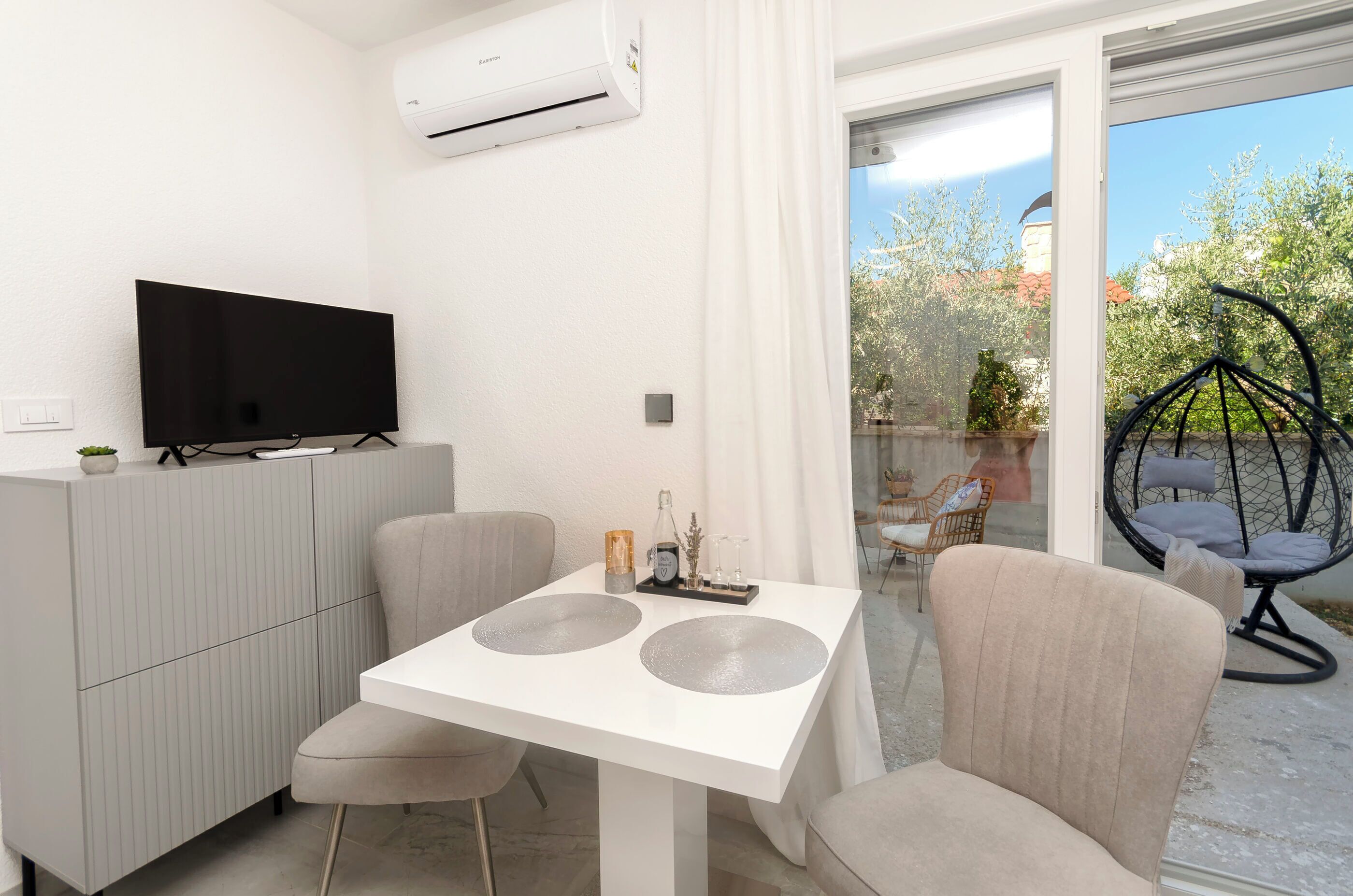 Una habitación moderna con un televisor de pantalla plana sobre un mueble, una mesa de comedor con dos sillas y vistas al exterior a través de una puerta de vidrio.