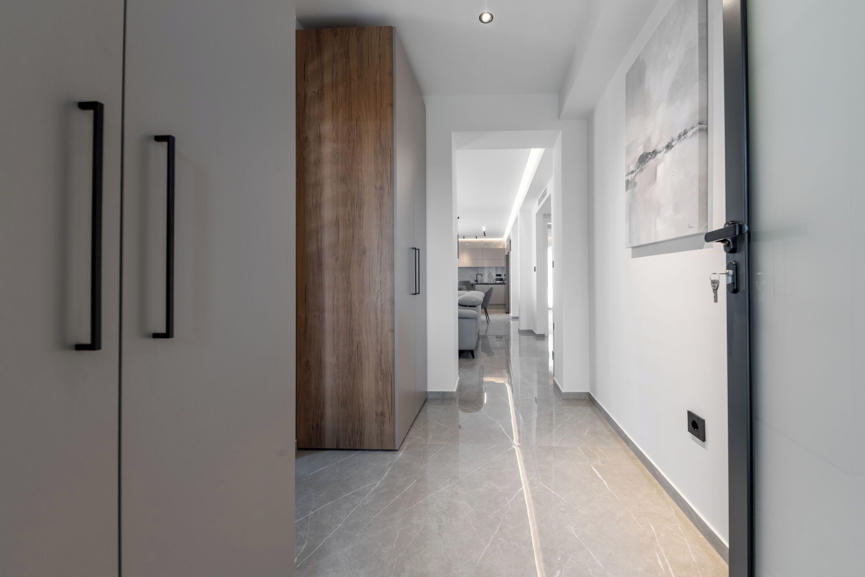 A modern hallway with a wooden door, a kitchen in the background, and a tiled floor.