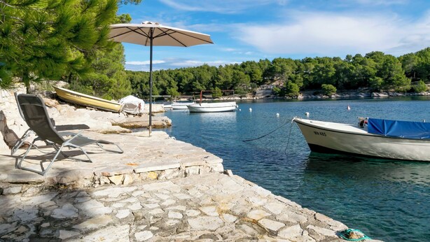 A stone pier with a lounge chair and umbrella, boats docked nearby, and a forested hillside in the background. - Apartments Jase (Milna)