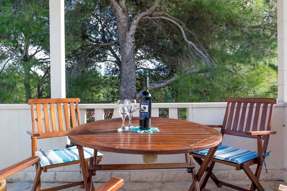 Une table en bois avec deux chaises, une bouteille de vin et deux verres, le tout sur un balcon avec un arbre en arrière-plan.