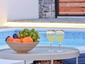 A table with a bowl of fruit and two glasses of a yellow beverage, near a pool. - Apartment Vransko (Pirovac)