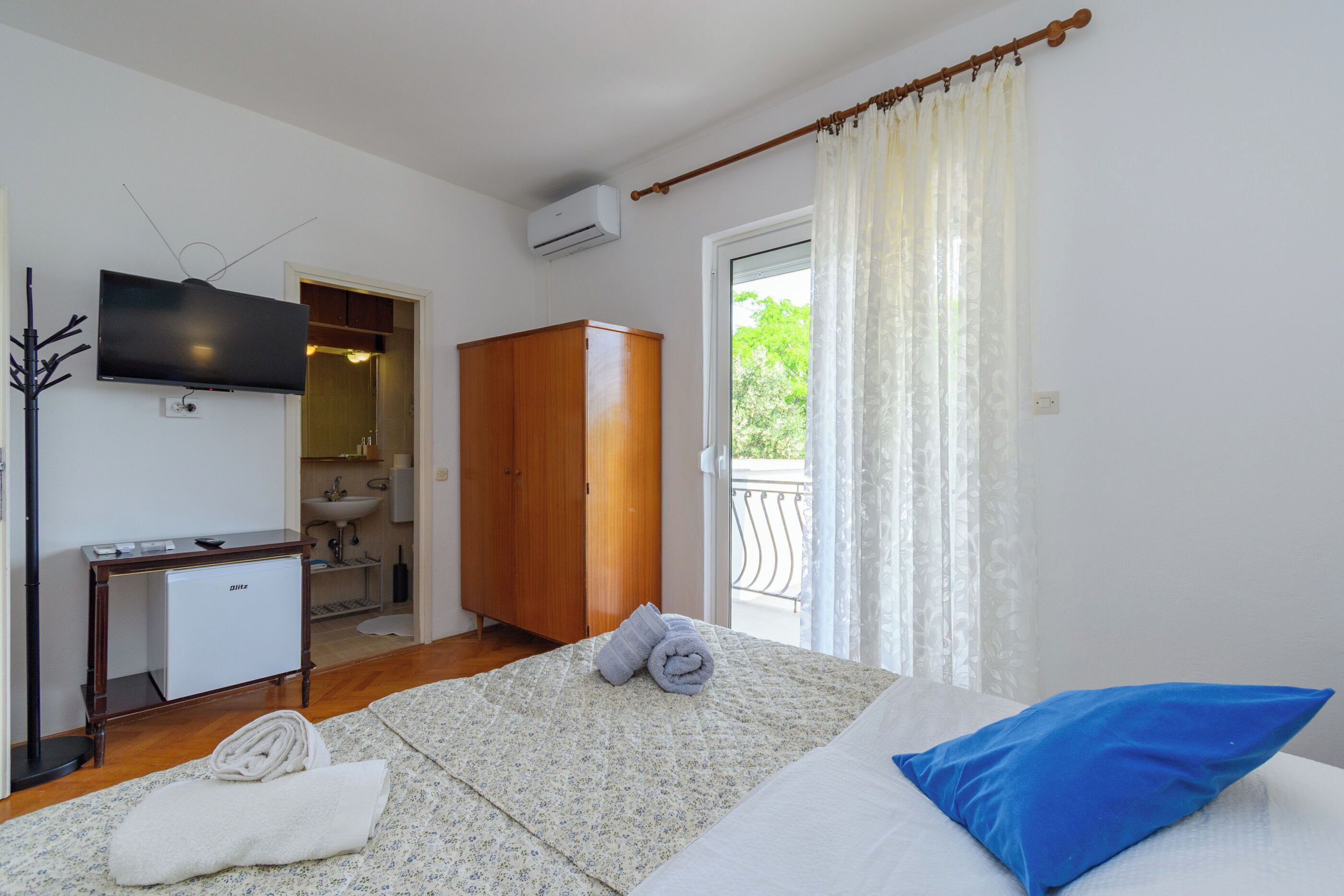 A bedroom with a bed, a TV, a wardrobe, a small desk, and a bathroom visible through an open door.