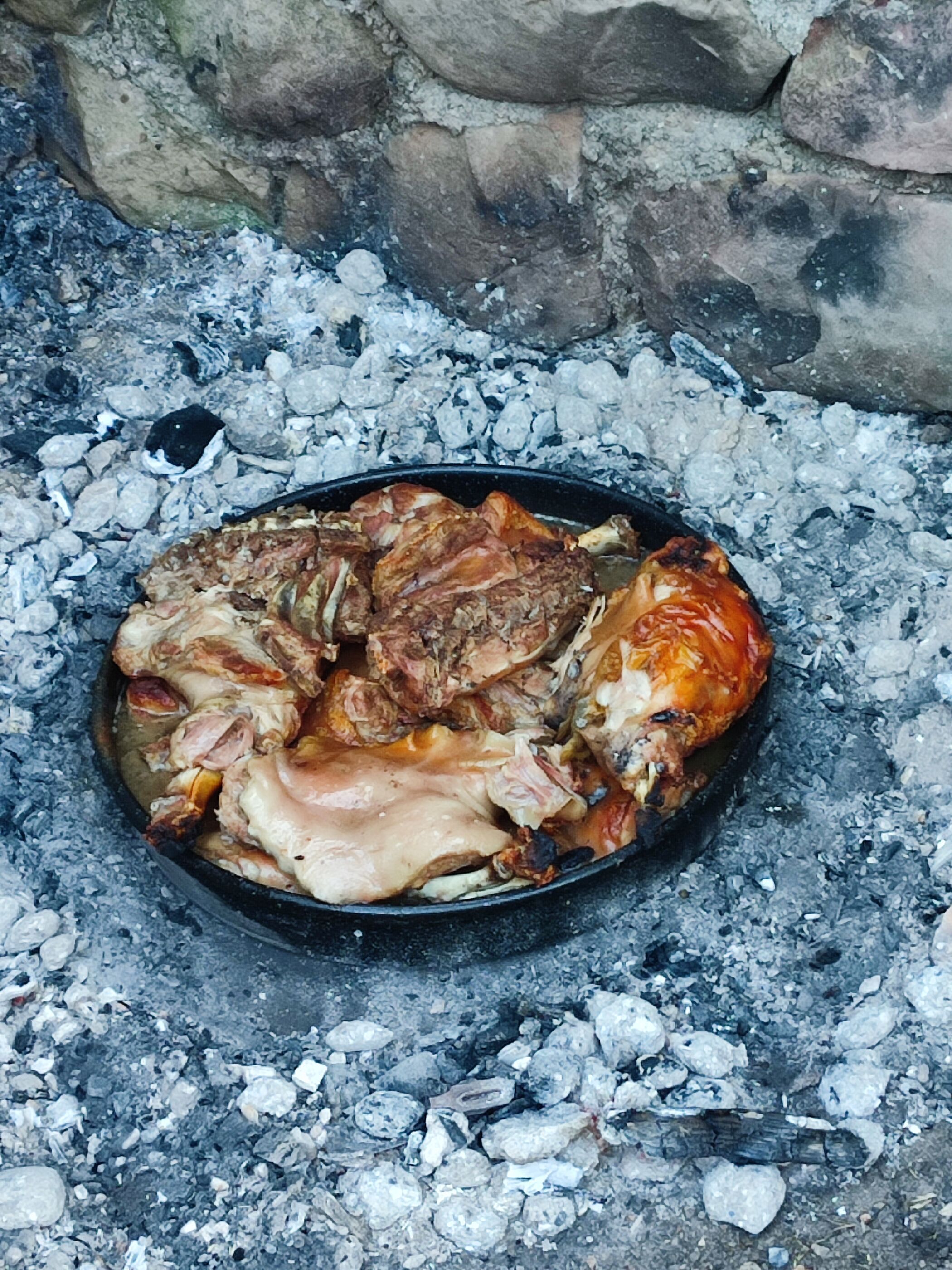 A round black cooking pot filled with roasted meat and vegetables on a bed of charcoal.