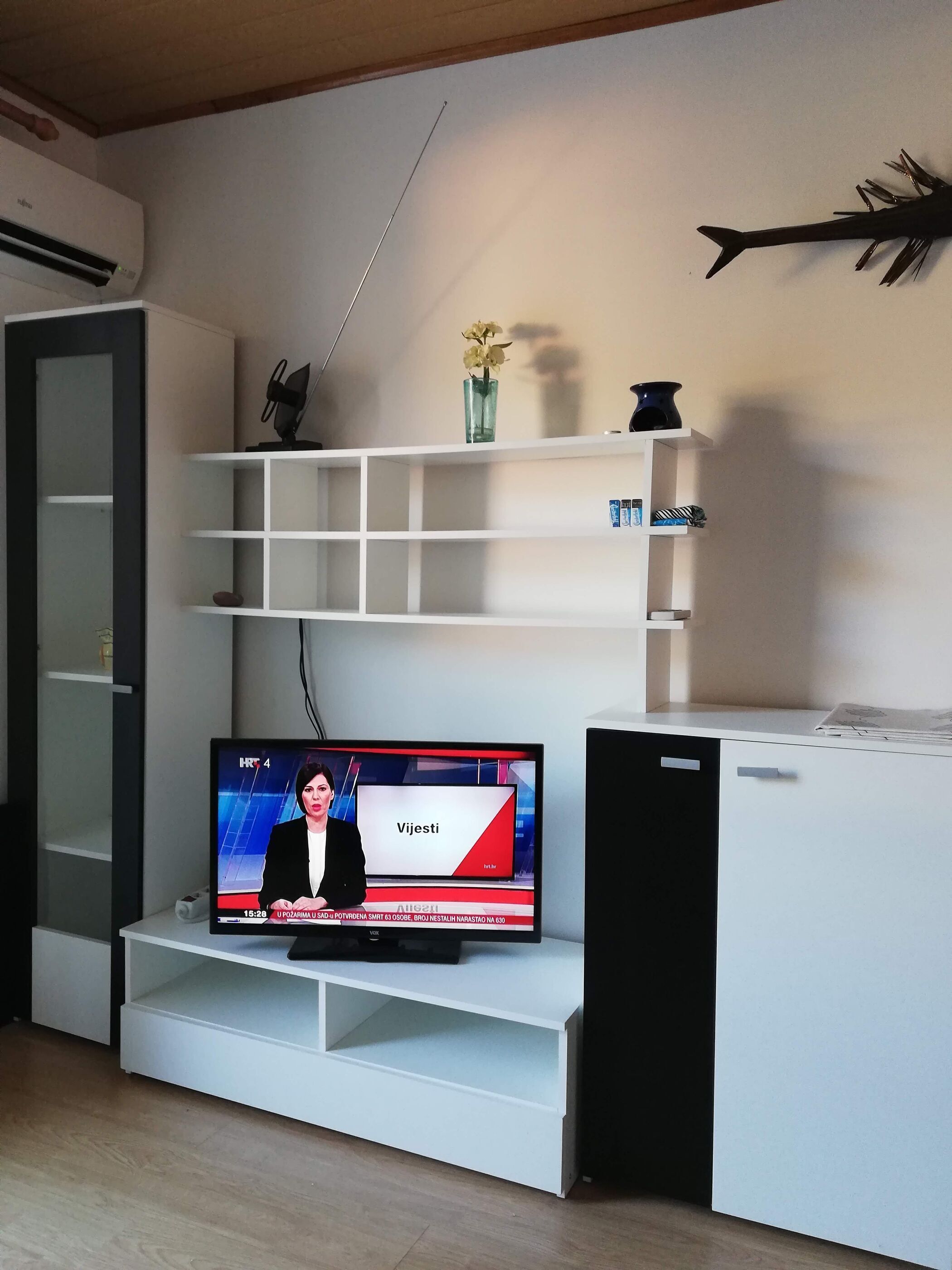 Ein modernes Wohnzimmer mit einem Flachbildfernseher auf einer weißen Unterhaltungseinheit, einer kleinen Ablagefläche mit Dekorationen und einer Tür links.