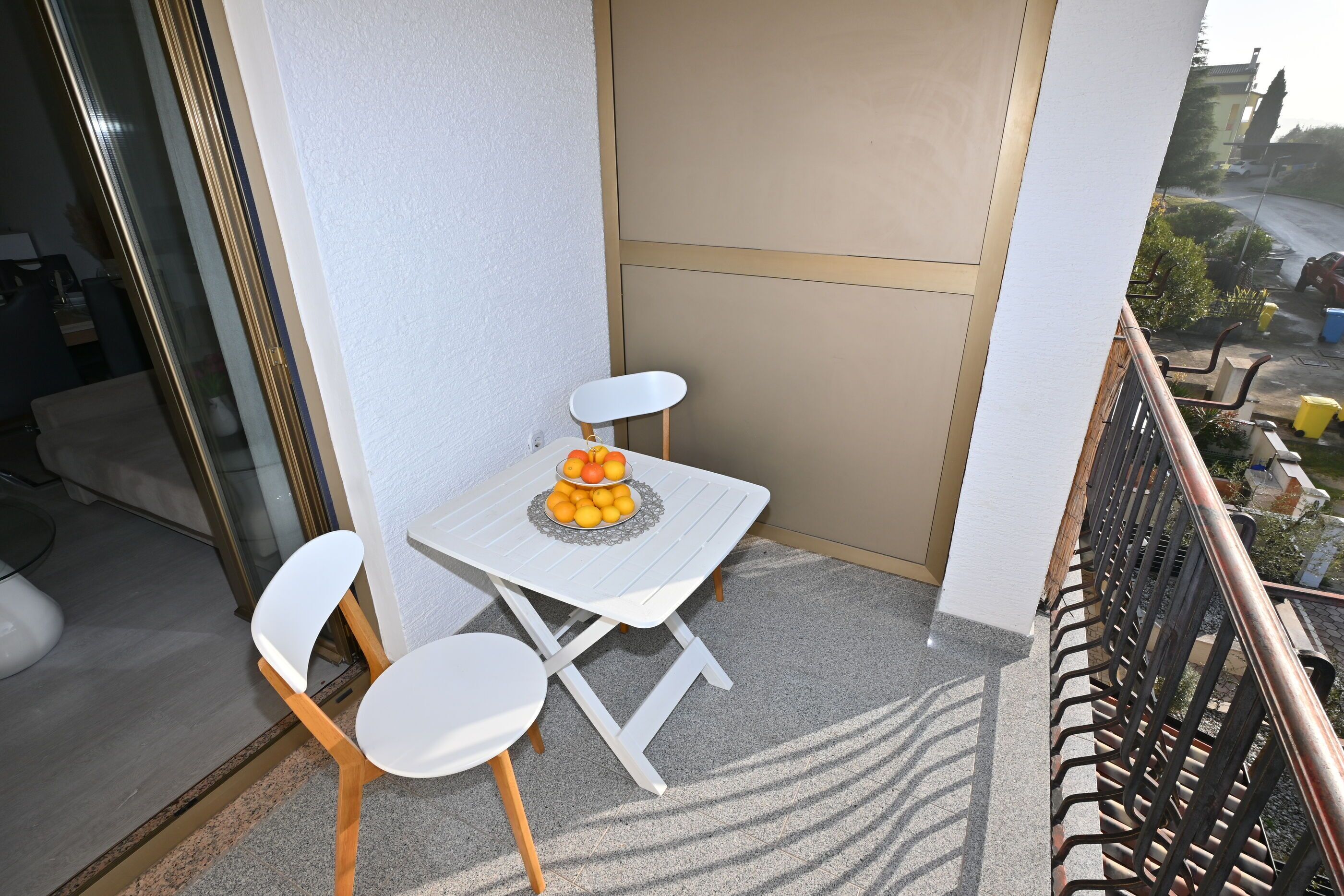 A small balcony with a white table and two white chairs, a fruit bowl on the table, and a view of the street below.