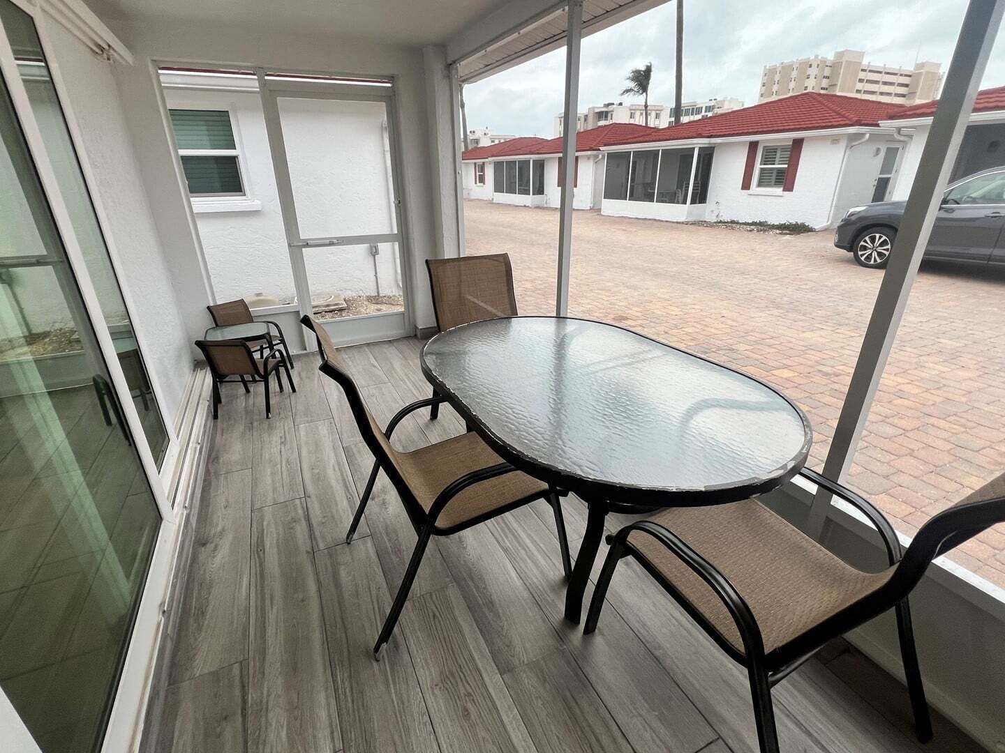 Une pièce avec une table et des chaises en verre, donnant sur un parking, et un bâtiment aux toits rouges en arrière-plan.
