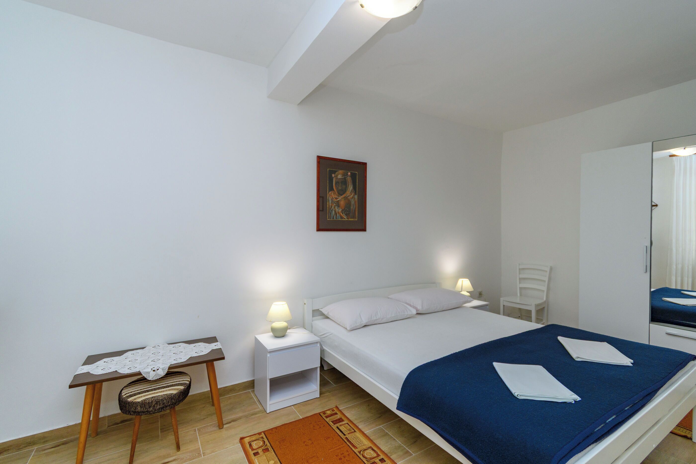 A bedroom with a bed, bedside tables, a chair, a small table with a lace tablecloth, a framed picture on the wall, and a mirror.