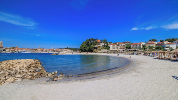 A beach with a rocky breakwater, clear blue water, and buildings in the background. - House Toni (Rab)