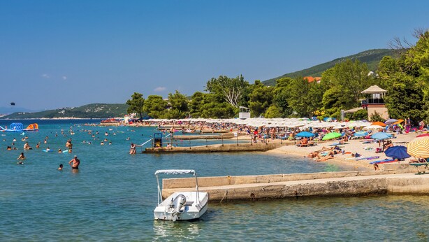 A beach with people swimming and sunbathing, a boat docked near the shore, and a lifeguard tower in the background. - Room Laura (Crikvenica)