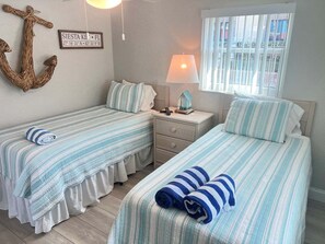 A bedroom with two beds, a nightstand, a lamp, a wall anchor, and a window with blinds. - Casa Blanca - Villa D02 (Siesta Key)