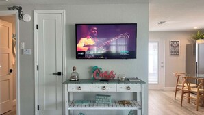 Un salon avec un téléviseur fixé au mur diffusant une vidéo musicale, un meuble de rangement blanc agrémenté d’objets décoratifs et une table à manger en bois.