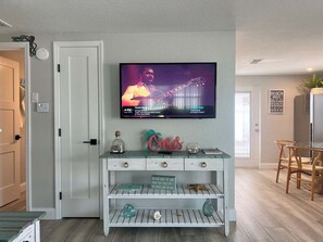 Ein Wohnzimmer mit an der Wand montiertem Fernseher, der ein Musikvideo zeigt, einer weißen Unterhaltungsanlage mit Dekorationsobjekten und einem Holzessttisch.