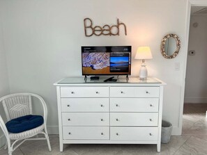 Eine weiße Kommode, ein Flachbildfernseher der eine Naturaufnahme zeigt, ein weißer Stuhl mit blauem Kissen und eine Wand mit dem Schild 'Beach'.
