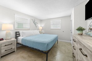 Ein Schlafzimmer mit einem Bett, Nachttischen, einem Schminktisch, einem an der Wand montierten Fernseher und einem Fenster mit Jalousien.