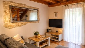 A living room with a stone wall, wooden beams, a grey sofa, a TV, and a window with sheer curtains. - House Ivana (Krsan)