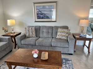 Ein Wohnzimmer mit grauem Sofa, zwei Beistelltischen mit Lampen, einem Holztisch mit Vase und einem gerahmten Bild an der Wand.