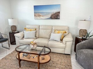 A living room with a beige sofa, two side tables, a glass coffee table, and a beach-themed painting on the wall. - Casa Blanca - Villa B16 (Sarasota)