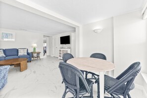 A modern living room with a sofa, coffee table, and wicker chairs around a central table. A flat-screen TV is mounted on the wall, and a door leads to another room. - Casa Blanca - Villa B14 (Sarasota)